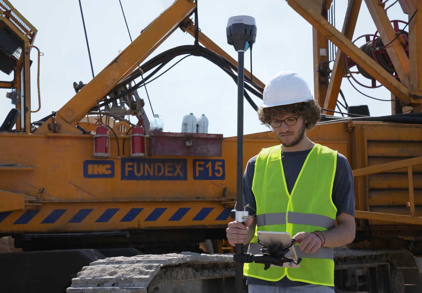 A surveyor checking grades on a tablet in front of an orange piling rig